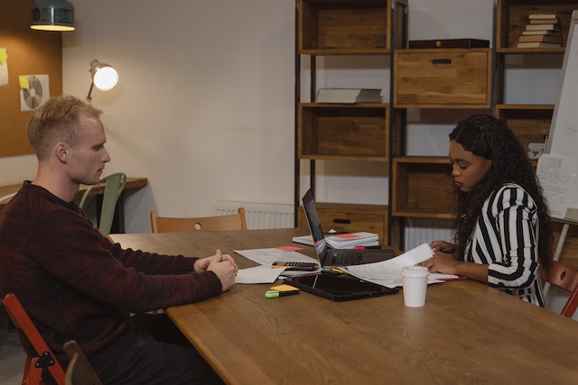 two people sitting across from one another with papers and a laptop between them